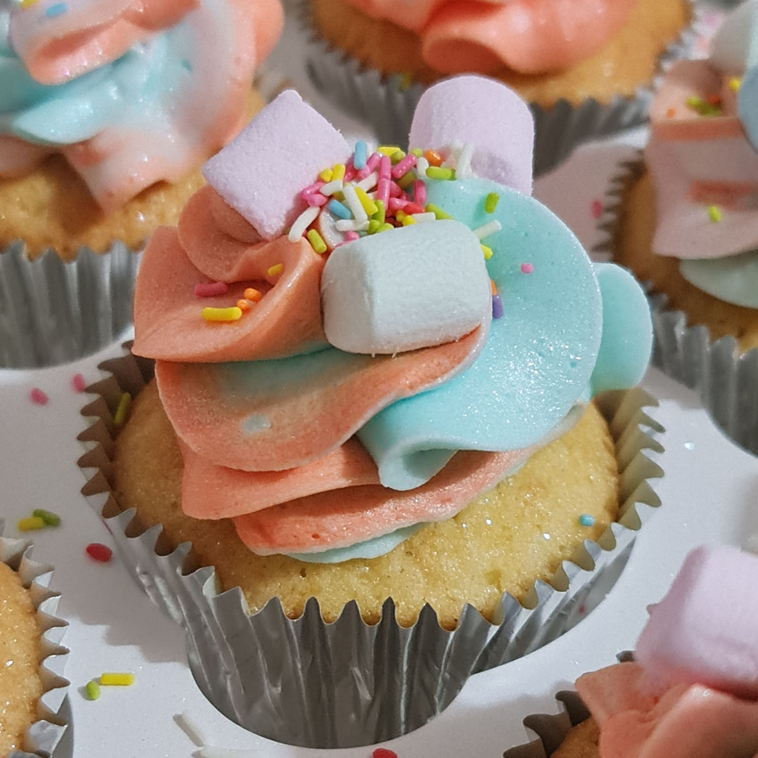 Beautiful light and fluffy cupcake with a light blue and light orange buttercream swirl on top with mini marshmallows and sprinkles sitting  between more cupcakes but focused mainly on the one,