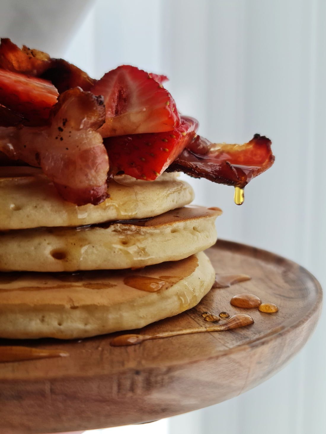 American fluffy pancakes topped with streaky bacon and strawberries. Drizzled in maple syrup on a wooden plate, on a white background